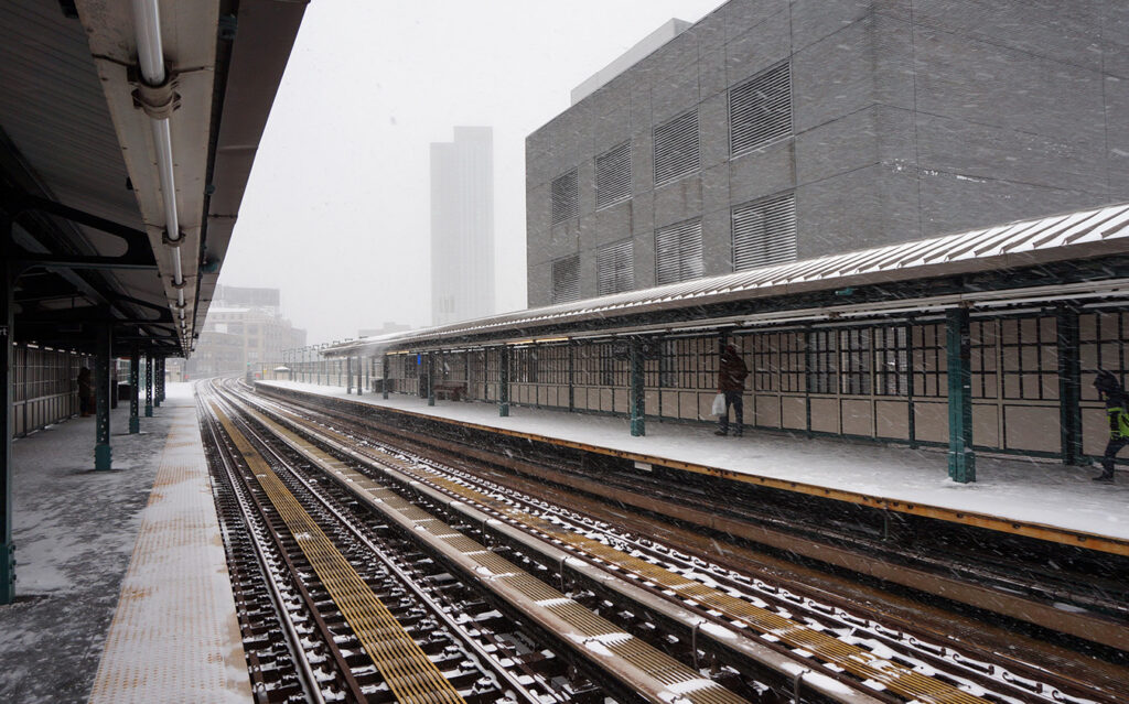 A snowy, empty elevated train station platform with tracks stretching into the distance; a few people stand under the covered area as snow falls and covers the ground, rooftops, and nearby townhouse facades.