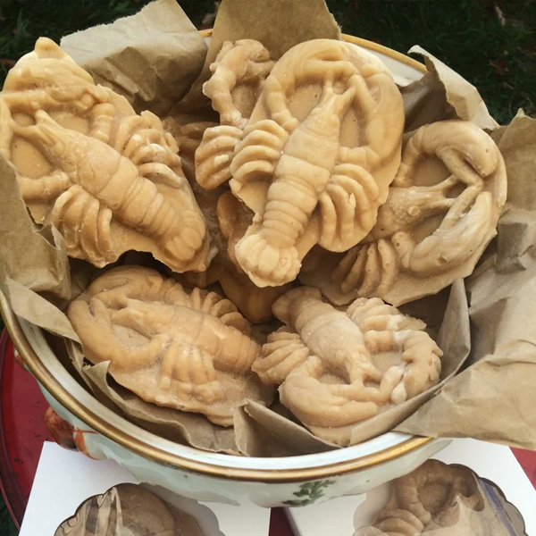 A bowl lined with brown paper holds several beige, molded candies shaped like lobsters—perfect for adding a whimsical touch to holiday decorating. The bowl is set outdoors on a surface with grass visible in the background.
