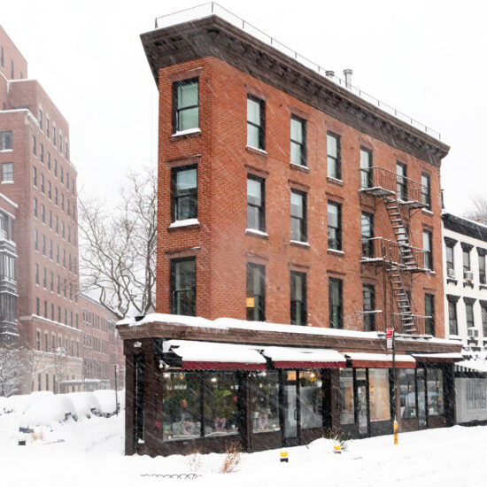 A red brick corner building with snowy rooftops and ground, featuring a fire escape and storefront windows, stands among other buildings on a city street during a heavy snowfall—a classic scene in the heart of Manhattan luxury sales.