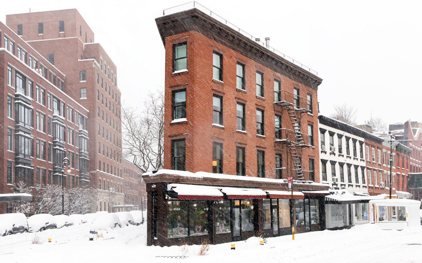 A red brick corner building with snowy rooftops and ground, featuring a fire escape and storefront windows, stands among other buildings on a city street during a heavy snowfall—a classic scene in the heart of Manhattan luxury sales.