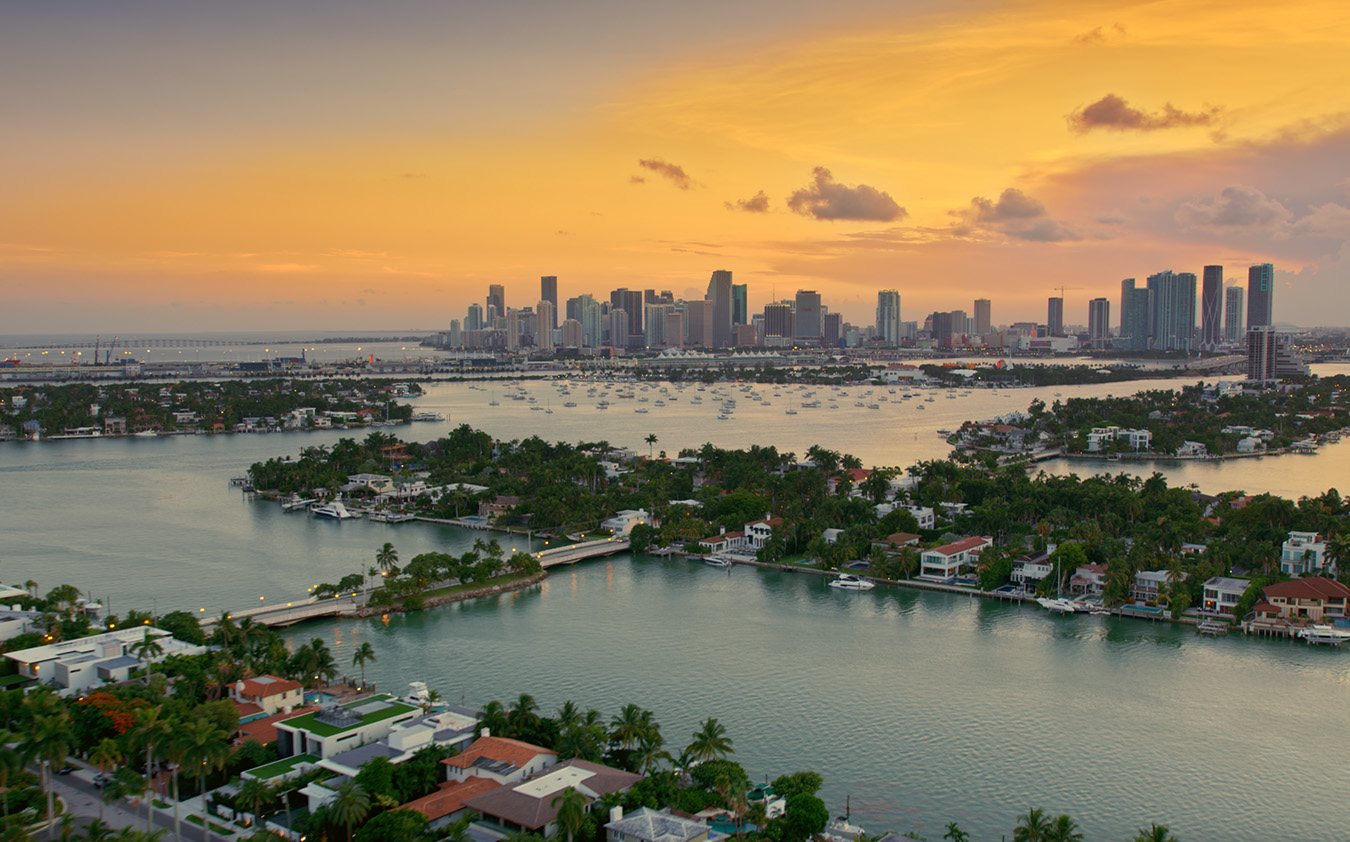 Aerial view of Miami at sunset, with the downtown skyline in the distance, surrounded by water, islands with houses, and boats scattered on the bay under a colorful sky.