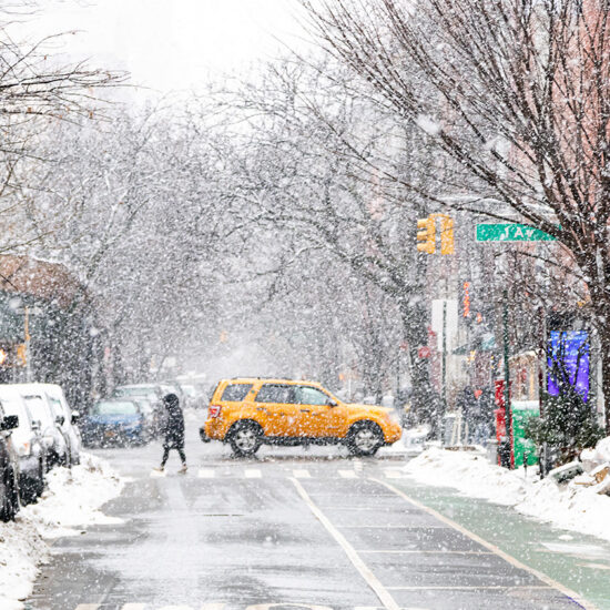 A yellow taxi drives through a snow-covered city street in an NYC residential rental neighborhood. Parked cars and trees line the street, while a person walks across the road. Snow blankets sidewalks and buildings in the background.