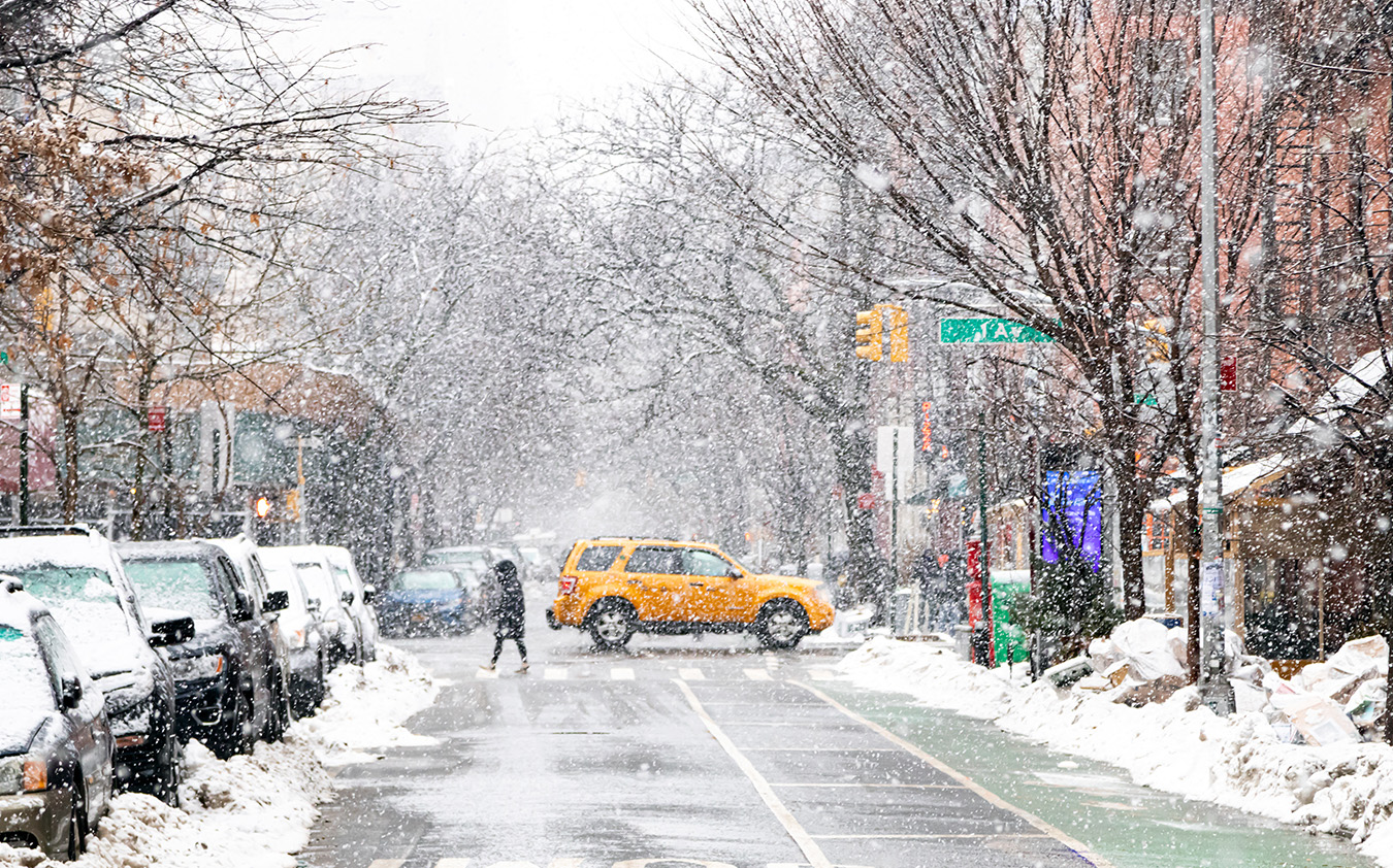 A yellow taxi drives through a snow-covered city street in an NYC residential rental neighborhood. Parked cars and trees line the street, while a person walks across the road. Snow blankets sidewalks and buildings in the background.
