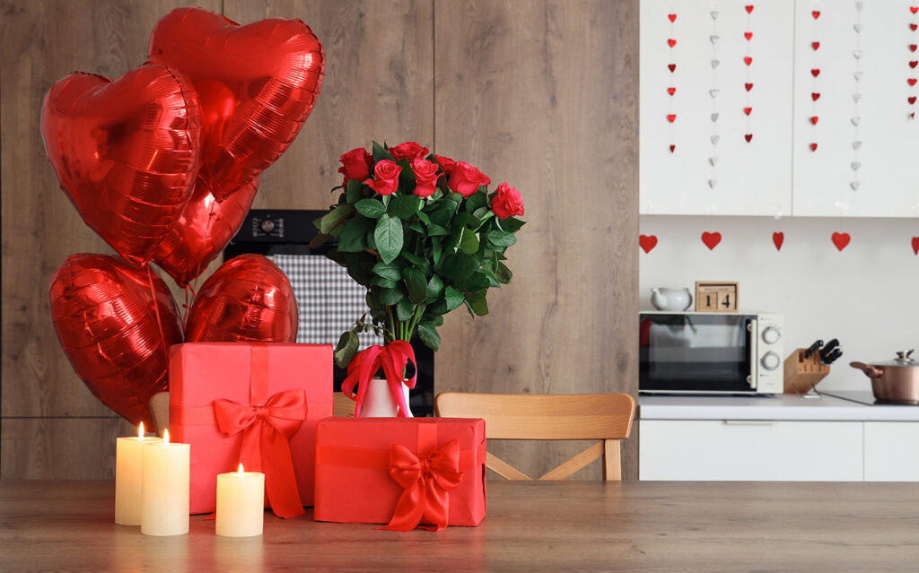 A table decorated for Valentine’s Day showcases holiday decorating with red heart-shaped balloons, a bouquet of red roses in a vase, two red gift boxes, and two lit candles. Heart decorations add festive charm to the kitchen background.