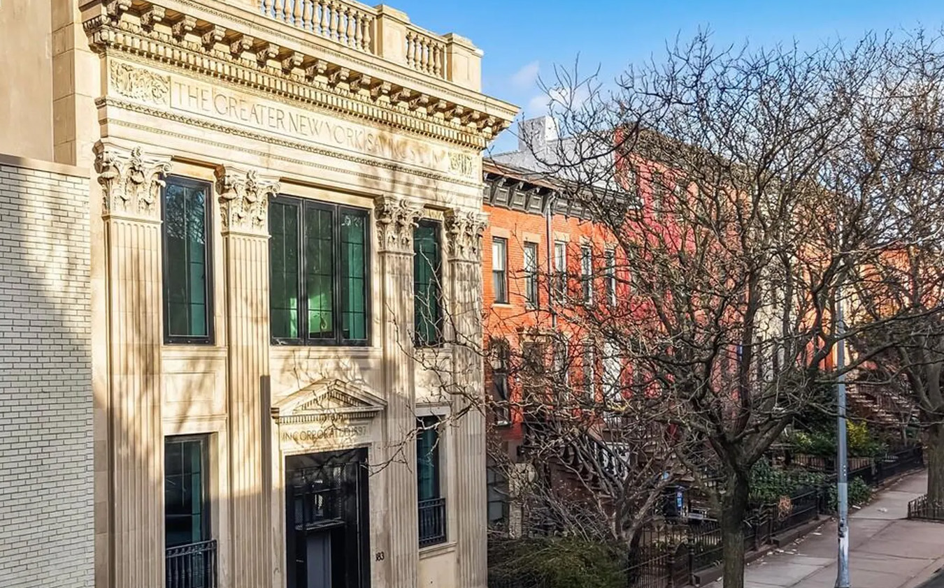 A historic beige stone building with ornate columns and “The Greater New York” engraved at the top stands on a sunny street lined with brick buildings and bare trees.