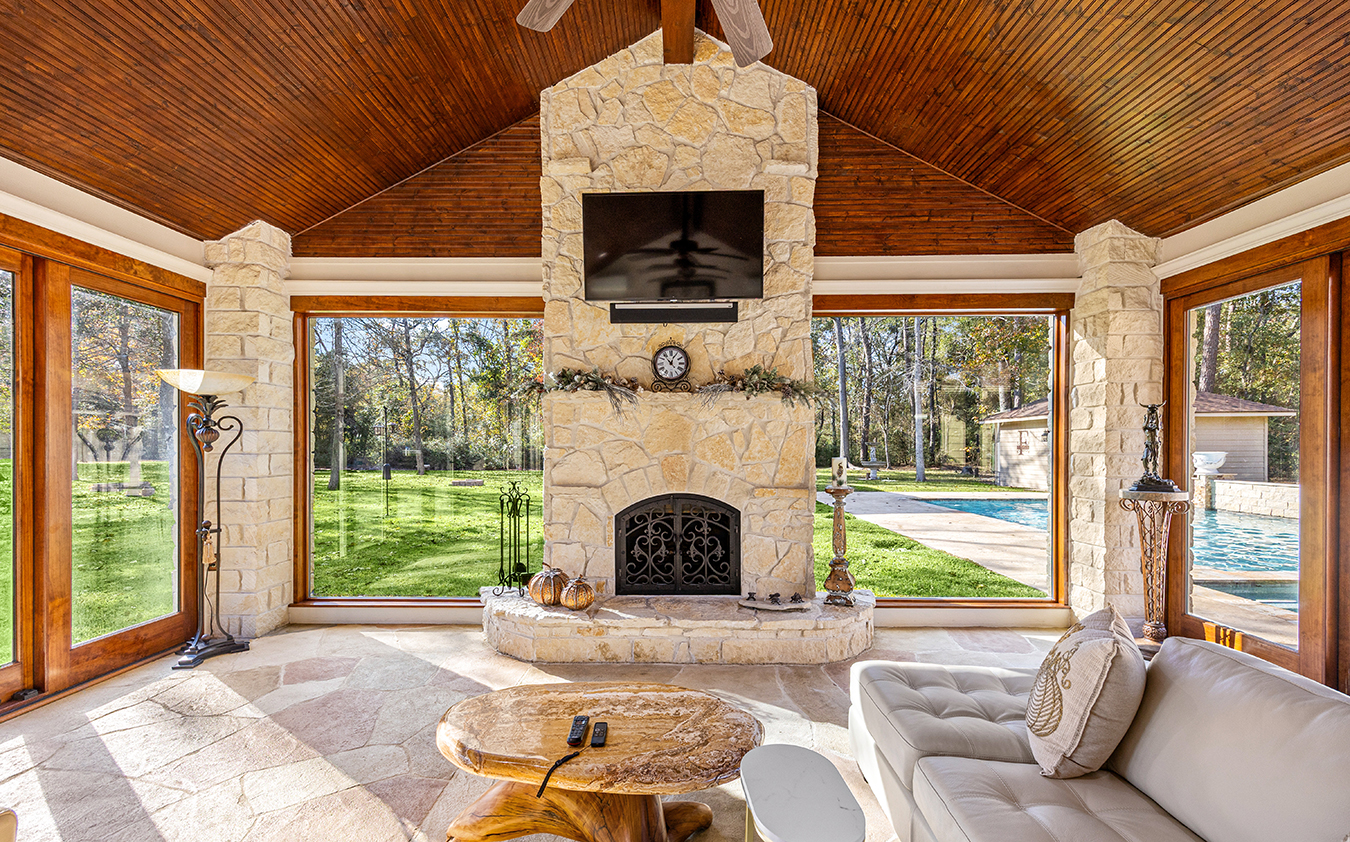 A bright sunroom with large windows, stone fireplace, mounted TV perfect for the big game, clock, beige sofa, round wooden table, and a view of a green lawn and swimming pool outside.