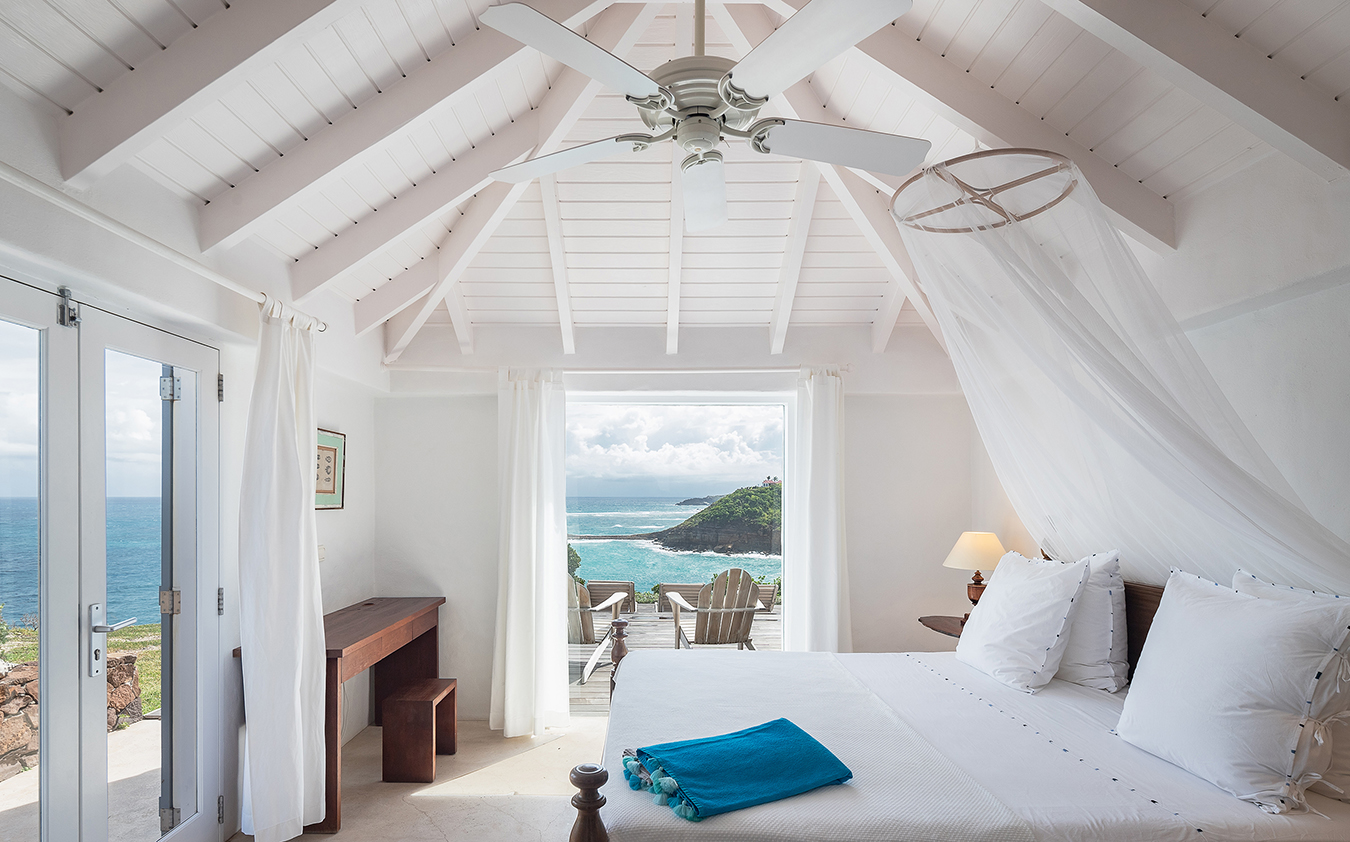A bright, airy primary suite with white walls and vaulted ceiling, featuring a bed with white linens, a blue towel, wooden furniture, and large doors opening to a balcony with ocean and cliff views.