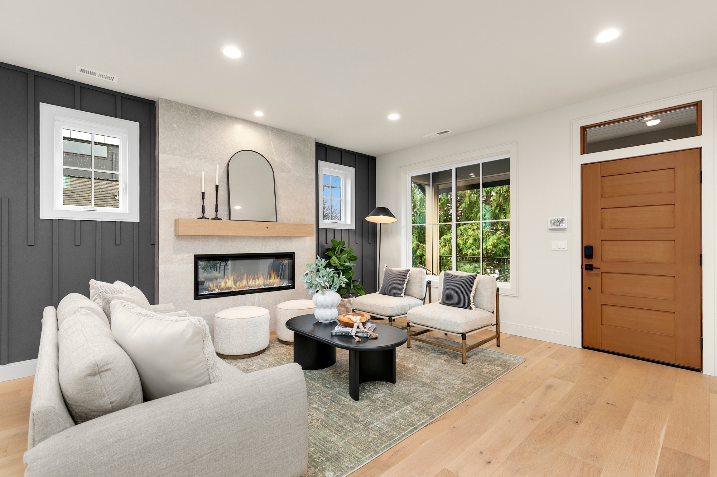 A modern living room with a gray sofa, two light chairs, and a black coffee table feels ready for an A-list: April 2026 gathering, featuring a fireplace under a mirror, large windows with greenery views, wood floors, and a wooden front door.