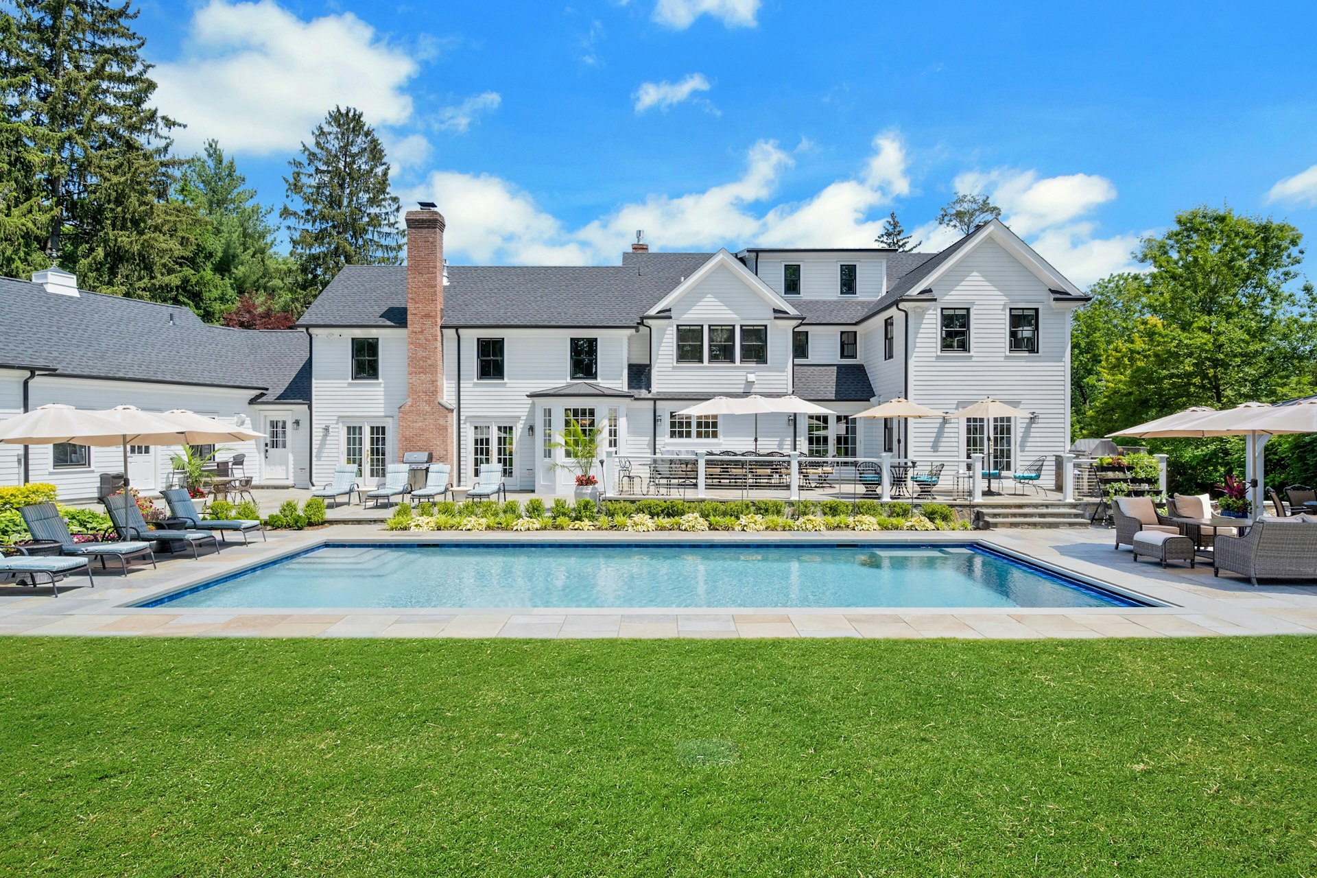 A large, modern white house with multiple windows and a brick chimney is shown behind a rectangular swimming pool, offering an a-list lifestyle with lounge chairs, umbrellas, and a manicured lawn under the bright April 2026 sky.