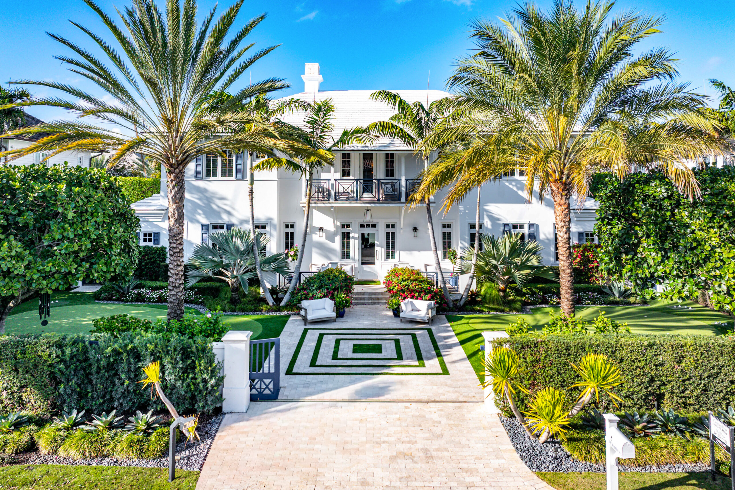 A large, white two-story house with a balcony is surrounded by palm trees, lush greenery, and manicured gardens. The geometric driveway leads to the entrance, framed by hedges and tropical plants—perfect for your A-list: April 2026 retreat.