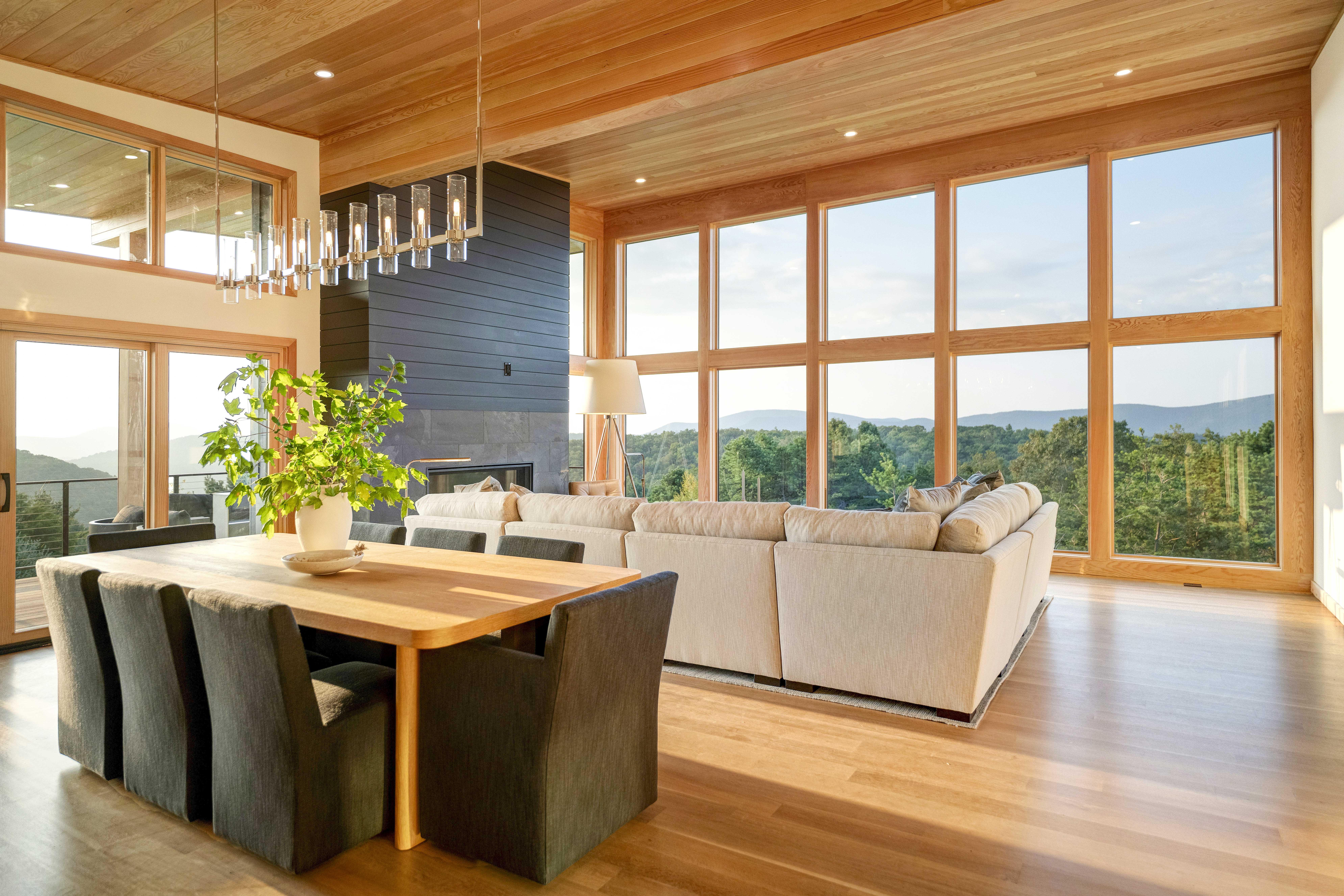 A modern living and dining area with large windows, wood flooring and ceiling, a light gray sectional sofa, black fireplace, dining table with black chairs, potted plant, and a scenic view of trees and hills—an a-list April 2026 retreat.