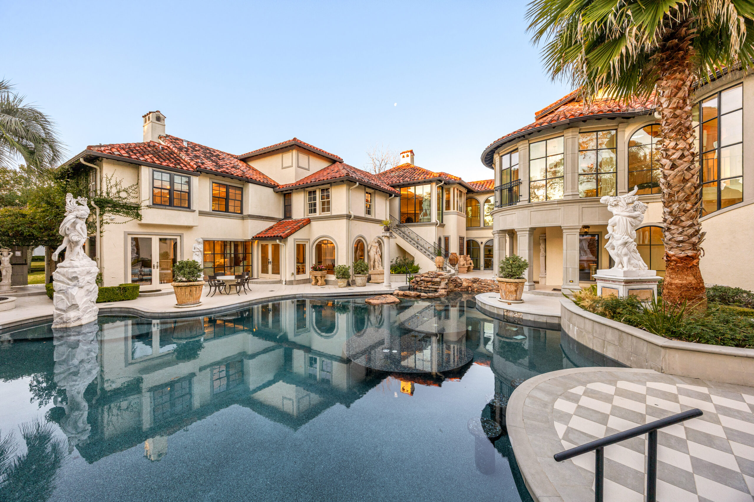 Large Mediterranean-style mansion with a red-tiled roof, arched windows, statues, and palm trees surrounds a stunning center courtyard pool—an A-list April 2026 retreat reflecting the house and blue sky.