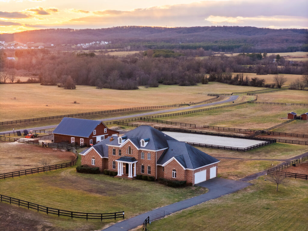 Aerial view of a large brick house with a circular driveway, surrounded by fenced fields, a barn, and an outdoor riding arena at sunset—perfect for those seeking prestigious equestrian estates in a serene rural landscape.