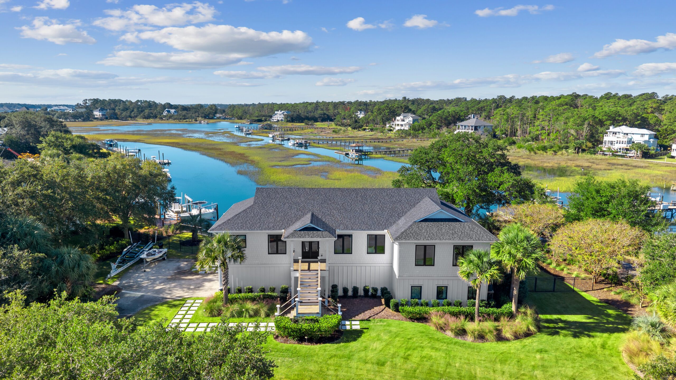 A modern two-story house with a gray roof sits by a scenic river, surrounded by trees, grass, and palm trees. Nearby docks and other homes complete this tranquil setting—an ideal retreat for an a-list April 2026 escape under blue skies.