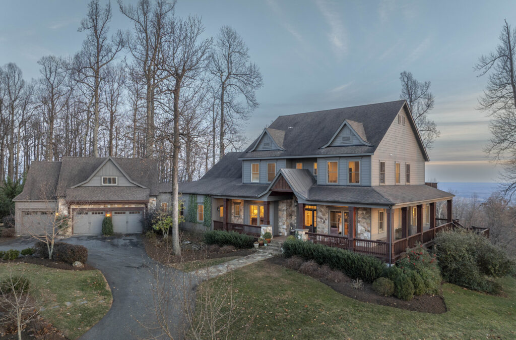 A large two-story house with a wraparound porch and a detached two-car garage sits among leafless trees at dusk. Warm lights glow inside, highlighting well-kept landscaping and a paved driveway—a serene retreat during Aries season.