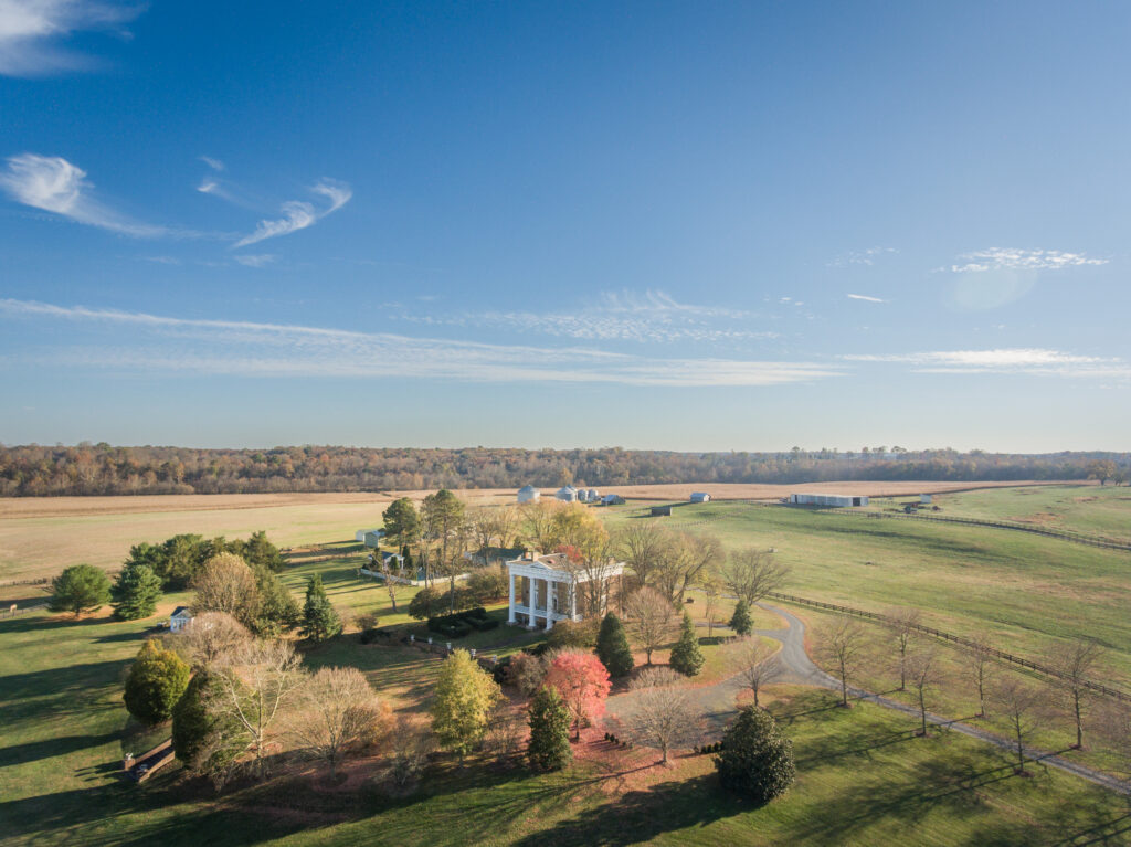 Aerial view of a large white plantation-style house surrounded by trees and open fields under a clear blue sky, with barns and farmland stretching into the distance—ideal for equestrian estates.