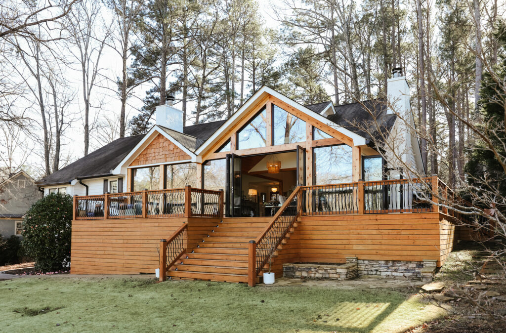 A modern house with large windows and a spacious wooden deck, set among tall trees—perfect for enjoying the energy of Aries season. The deck has stairs and railings, leading down to a grassy yard bordered by stone landscaping.