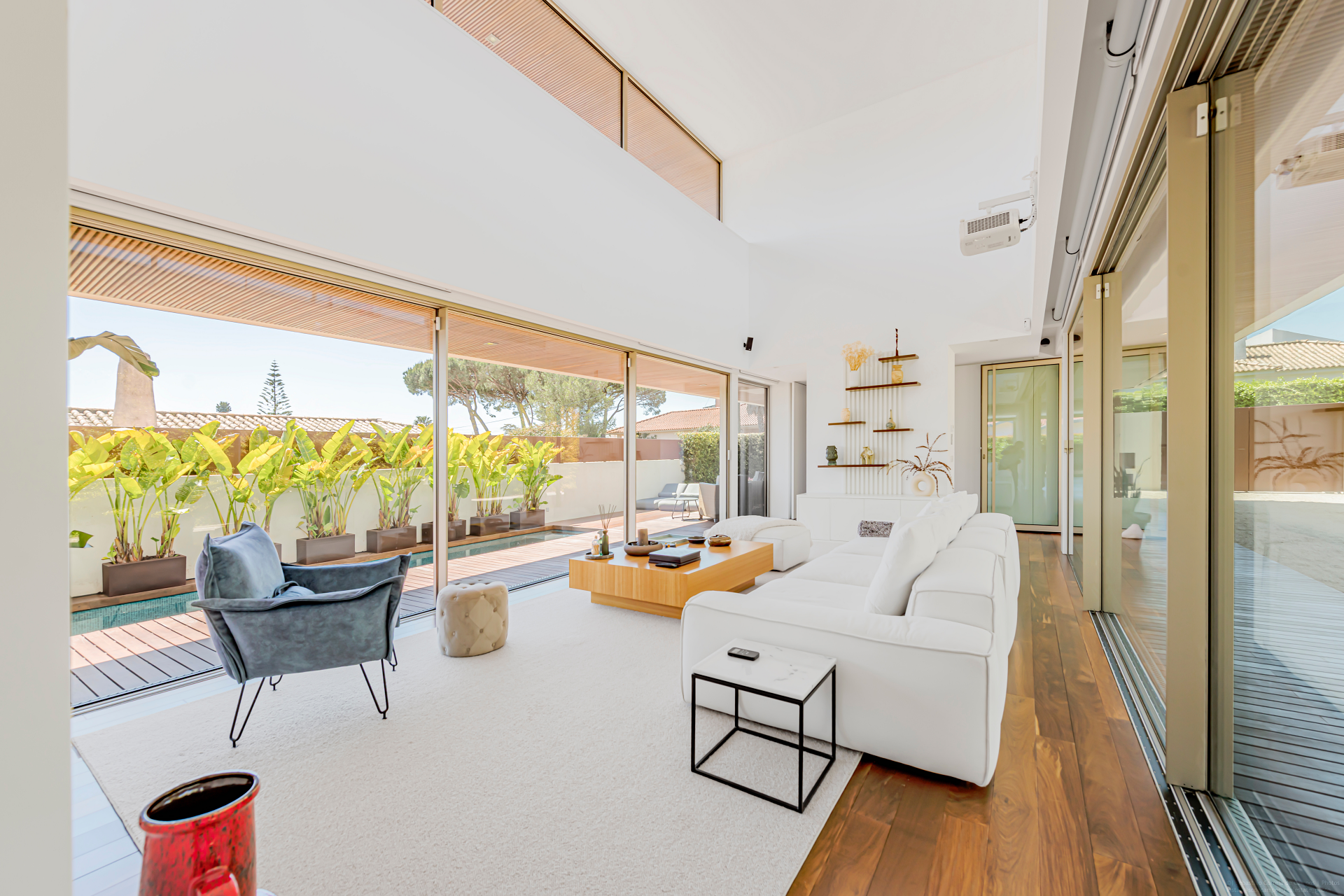 Bright modern living room with white sofa, glass walls, wooden floor, and potted plants outside. Space is filled with natural light and minimalist decor—an a-list retreat for April 2026, offering a cozy open atmosphere overlooking the garden patio.