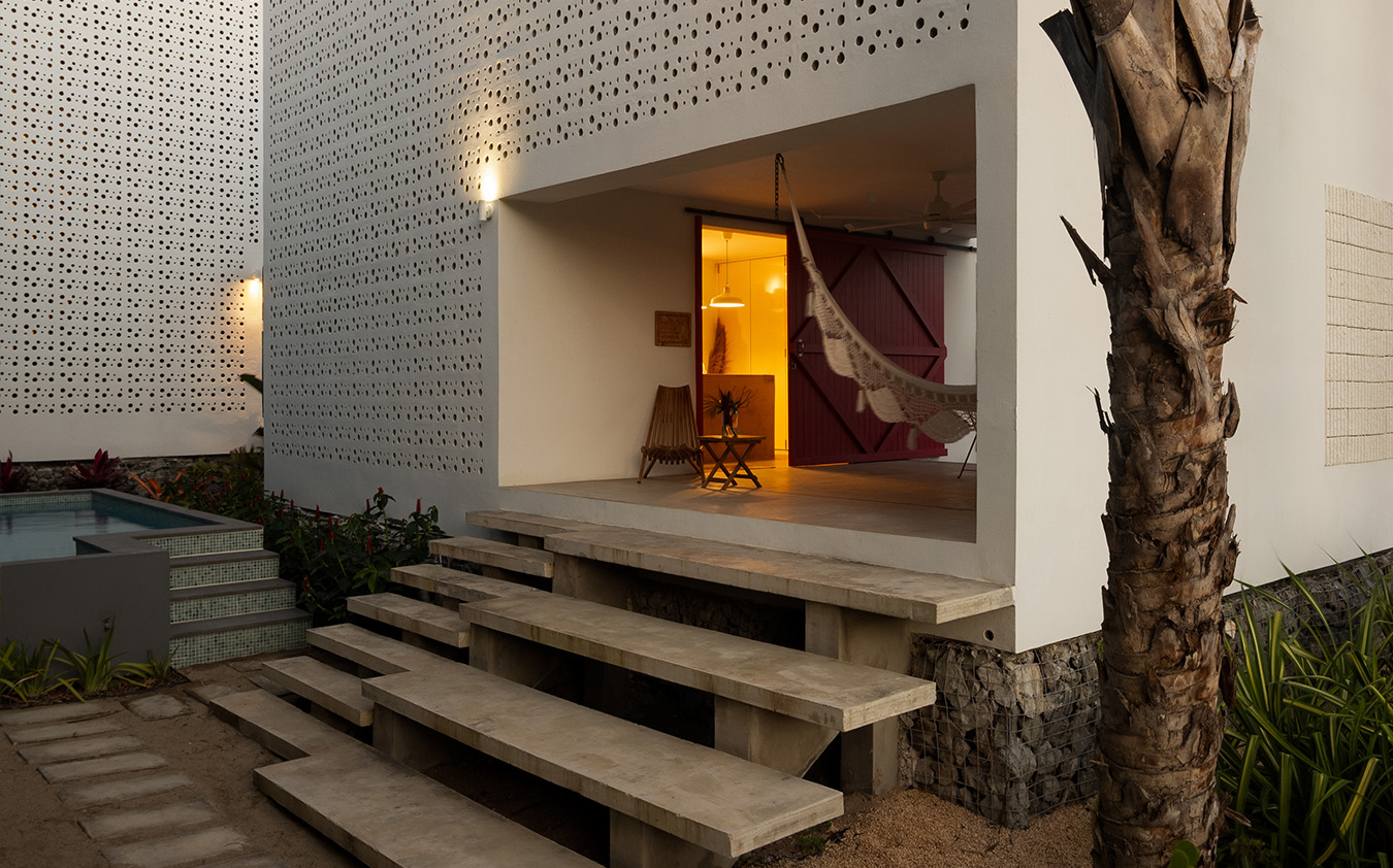 Modern house with perforated white walls, a wide concrete staircase leading to a porch with a hammock, wooden chair, and soft lighting. Featured on the A-List: April 2026. A small pool and palm tree are visible in the landscaped garden.