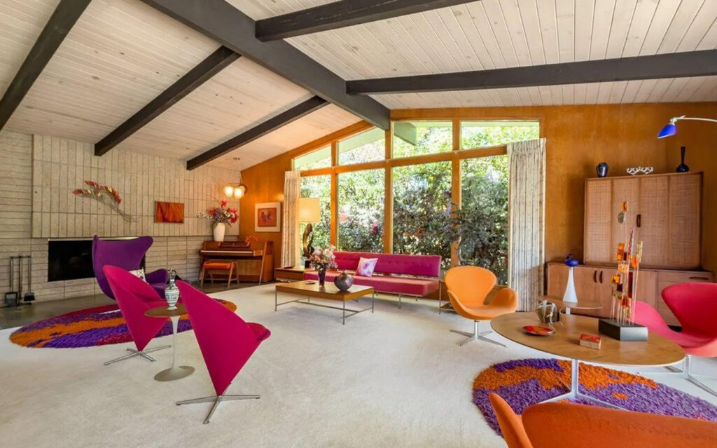 A retro living room with vaulted ceilings, exposed beams, large windows, colorful mid-century modern furniture in pink, orange, and purple—perfect for equestrian estates—plus a white brick fireplace, vibrant rugs, and vintage decor.