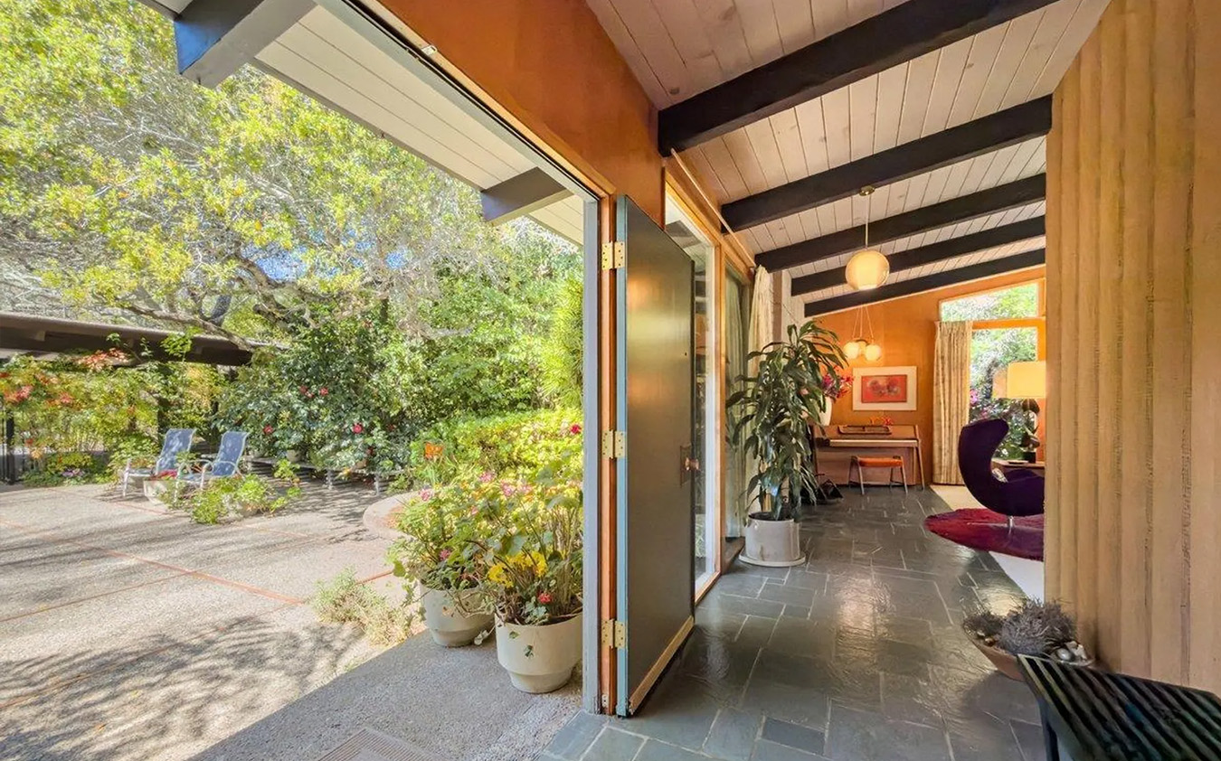 A bright mid-century modern home interior with a slate floor opens to a patio with potted plants and outdoor chairs, surrounded by lush greenery and trees—reminiscent of tranquil equestrian estates visible through large glass doors.