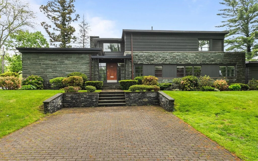 A modern two-story house with dark stone exterior, flat roof, large windows, and neatly trimmed bushes, set back from a brick driveway with stone steps and lush green lawn—perfect for those seeking refined living in equestrian estates.