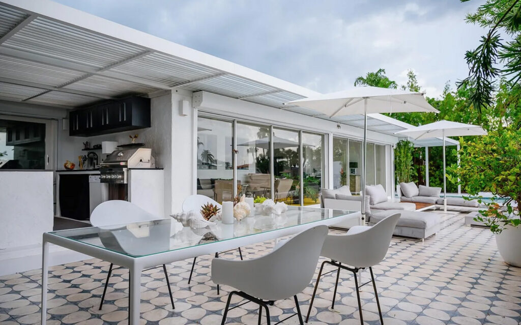 Modern outdoor patio in equestrian estates with a glass dining table and white chairs on patterned tile, lounge chairs with white umbrellas, greenery, and large glass doors leading inside. Grill and cabinetry are visible in the background.