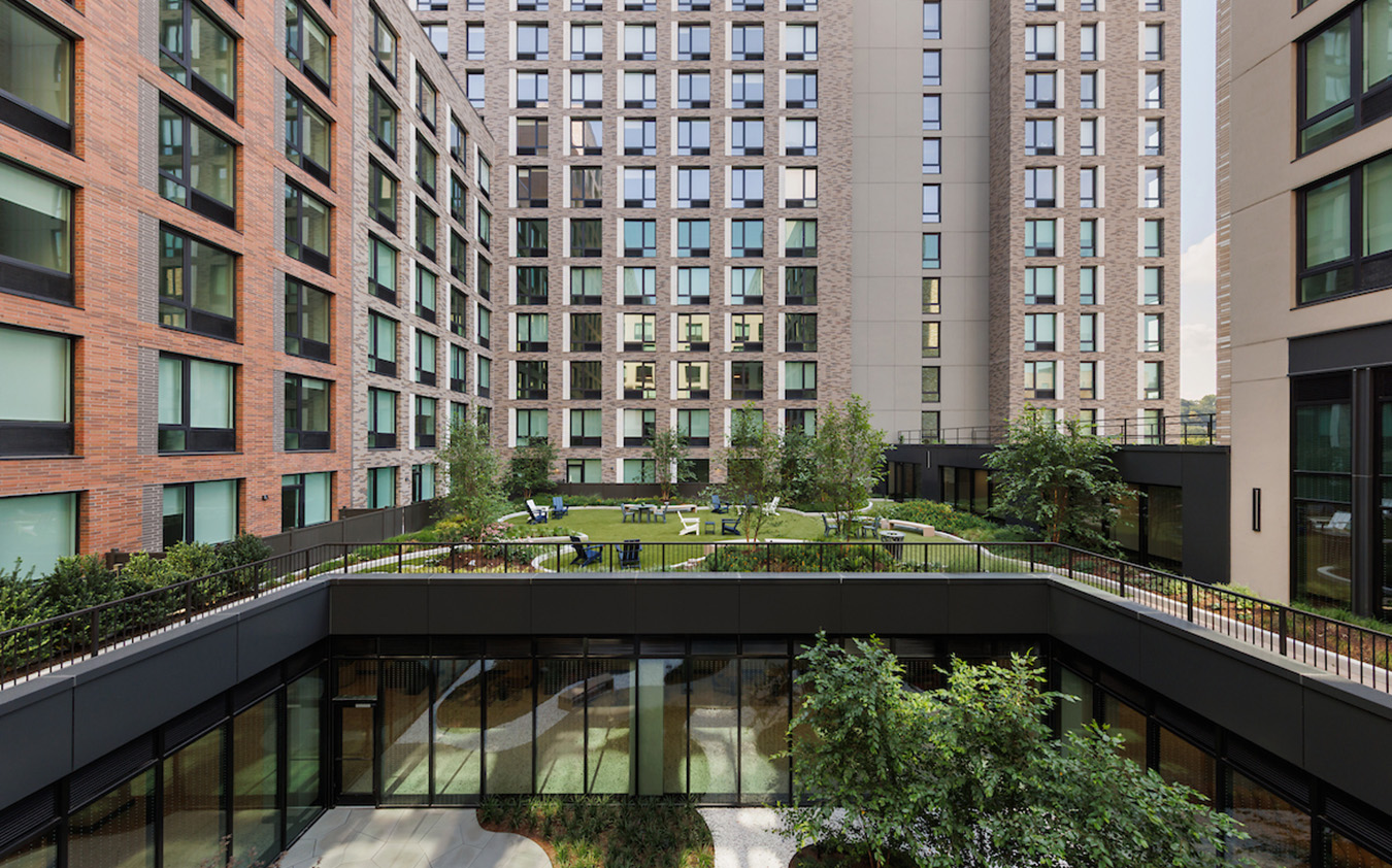 A modern apartment complex surrounds a landscaped courtyard with green grass, trees, and outdoor seating. Large windows line the surrounding buildings, reflecting the outdoor space.
