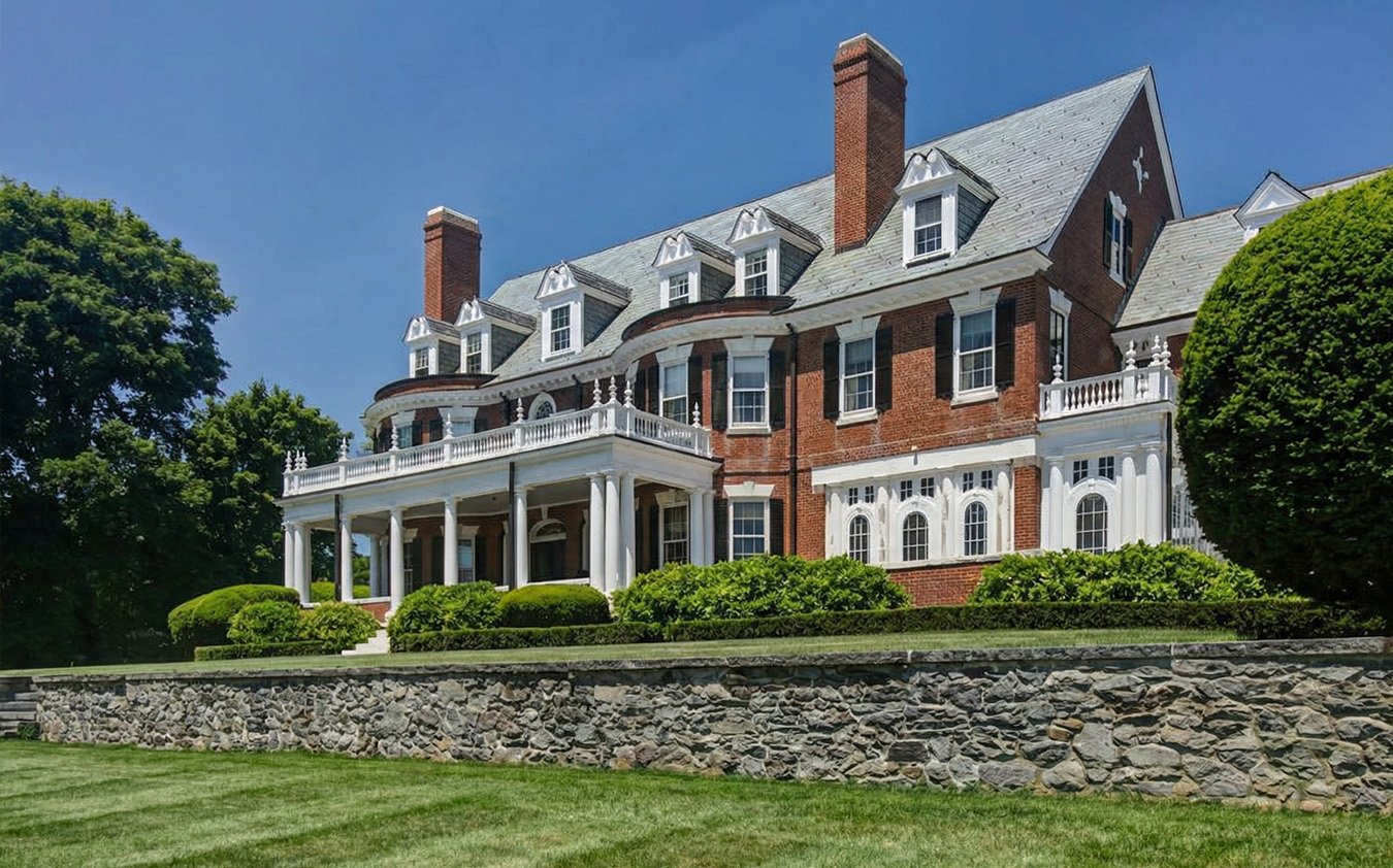 A large, elegant red-brick mansion with white trim, multiple chimneys, dormer windows, and a grand columned porch, set behind a stone wall and surrounded by manicured green lawns and bushes.