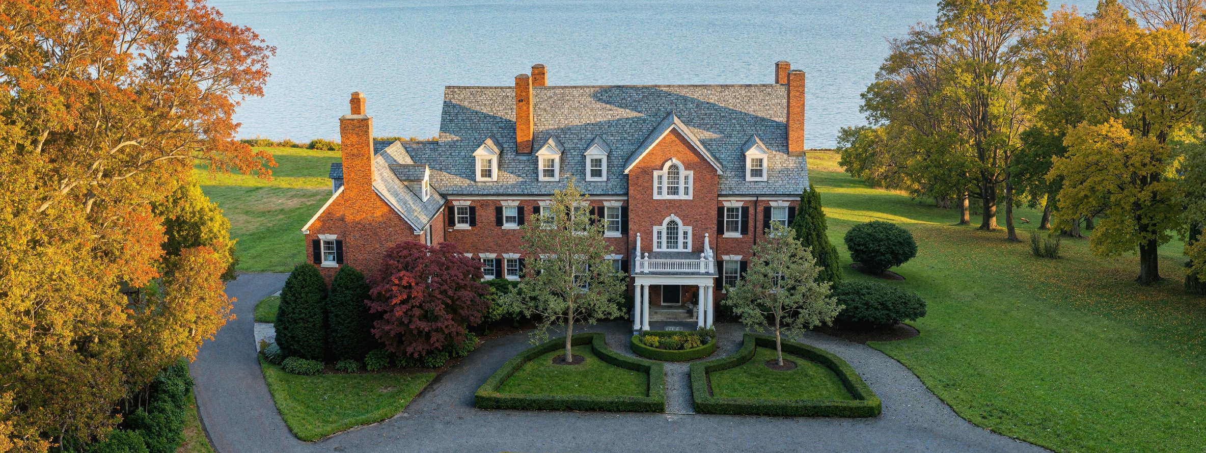 A large, red-brick mansion with white trim, multiple chimneys, and dormer windows sits amid manicured lawns and trees, overlooking a large body of water in the background.