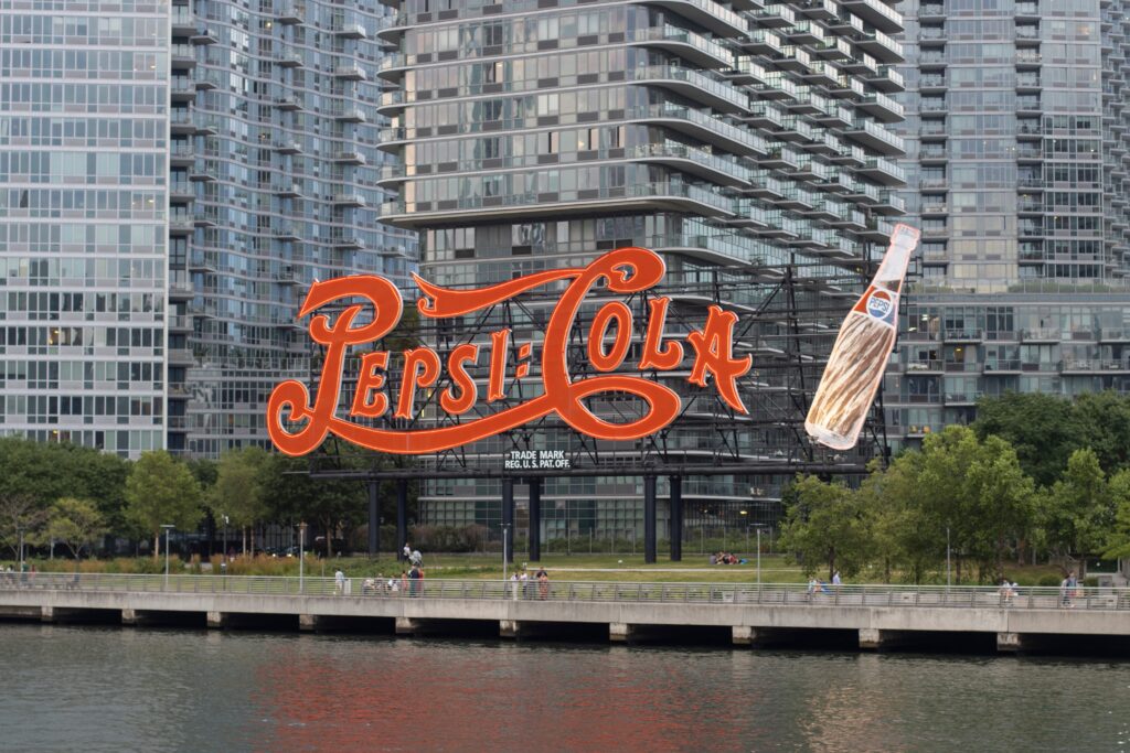 Pepsi-Cola Sign A large vintage Pepsi-Cola sign and a bottle sculpture—far from ordinary landmarks—stand in front of modern glass apartment buildings along a waterfront lined with trees and a scenic walkway.