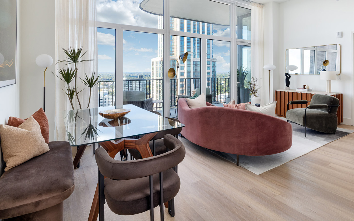 Modern living and dining area in a high-rise apartment with large windows, city views, glass dining table, burgundy sofa, green armchair, wooden floors, and potted plants.