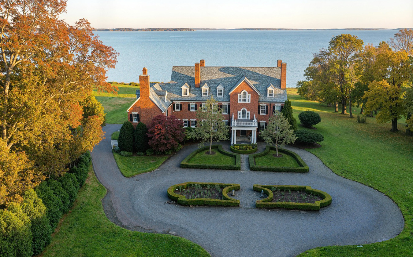 A large brick mansion with a circular driveway, manicured gardens, and surrounding green lawns sits near a calm body of water, with trees showing autumn colors in the background.