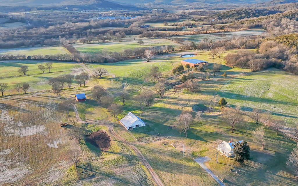 Aerial view of a rural landscape featuring scattered houses, barns, equestrian estates, green fields, trees, and winding dirt roads, set against hills beneath a partly cloudy sky.