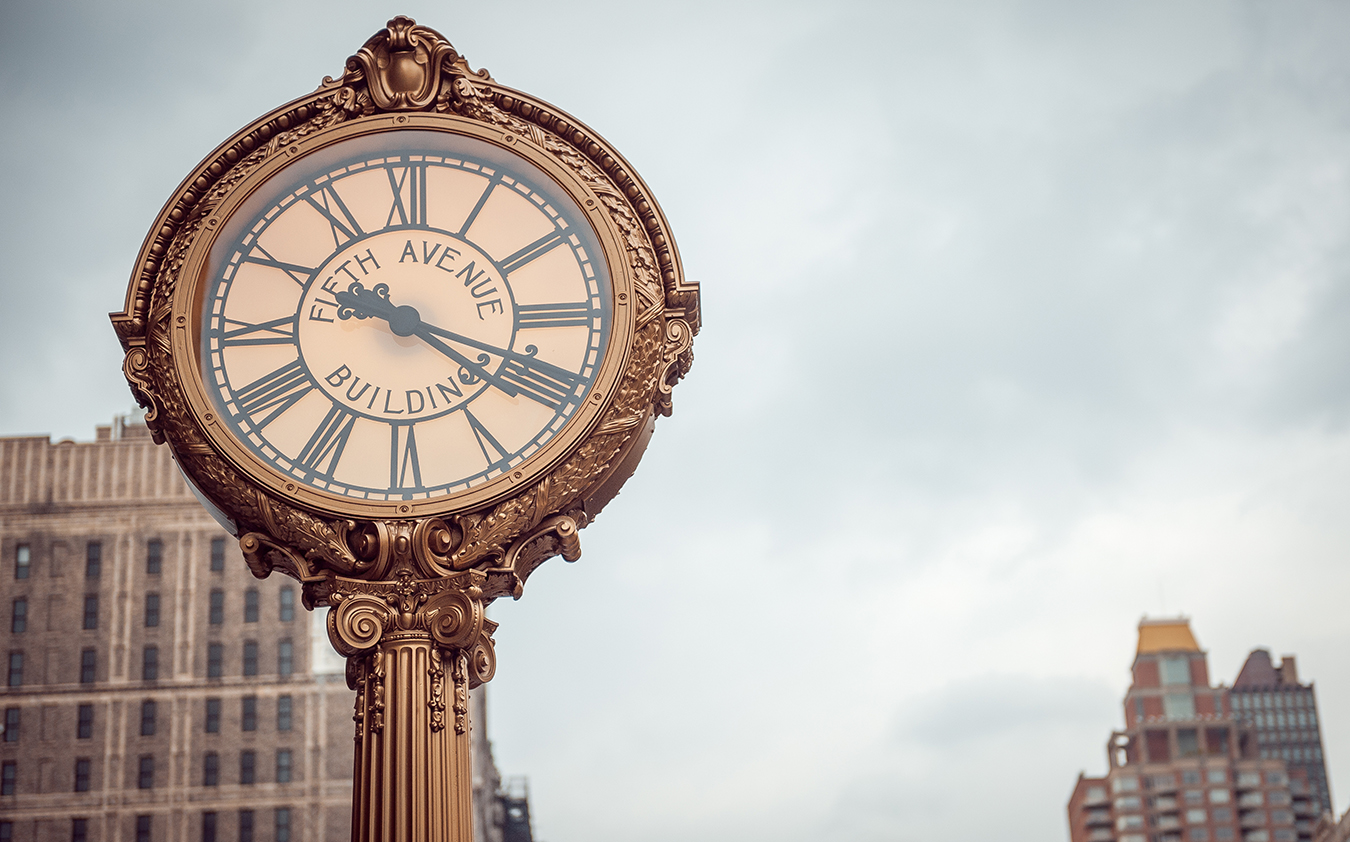 An ornate street clock with Fifth Avenue Building on its face, displaying Roman numerals, stands in front of tall buildings under a cloudy sky, elevating the charm of ordinary landmarks in an urban setting.