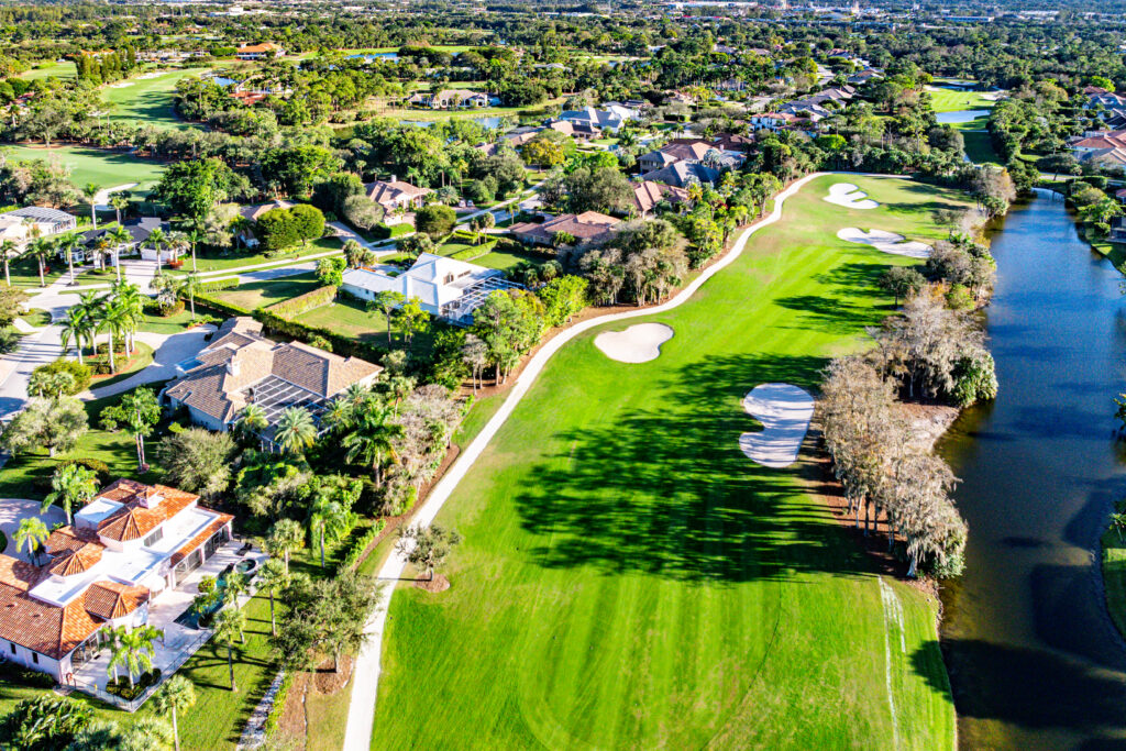 Aerial view of a suburban neighborhood with large houses bordering a golf course, featurin g sand bunkers, lush green fairways, a water hazard, and landscaped areas dotted with trees.
