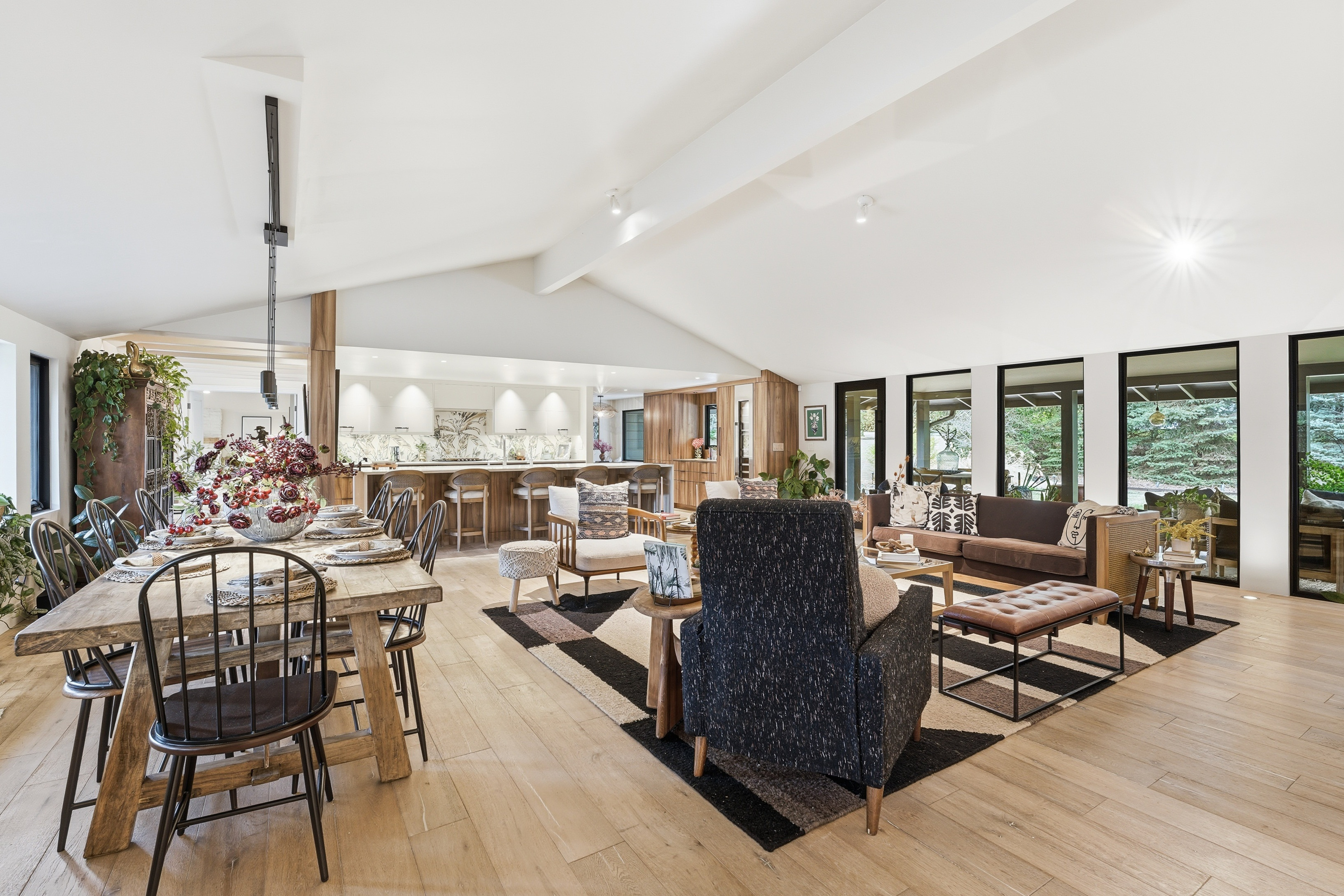Open-concept living and dining area with wooden floors, a rustic dining table with black chairs, cozy seating, large windows, and a modern kitchen in the background—an A-list space designed for comfort and style. Natural light fills the spacious room.