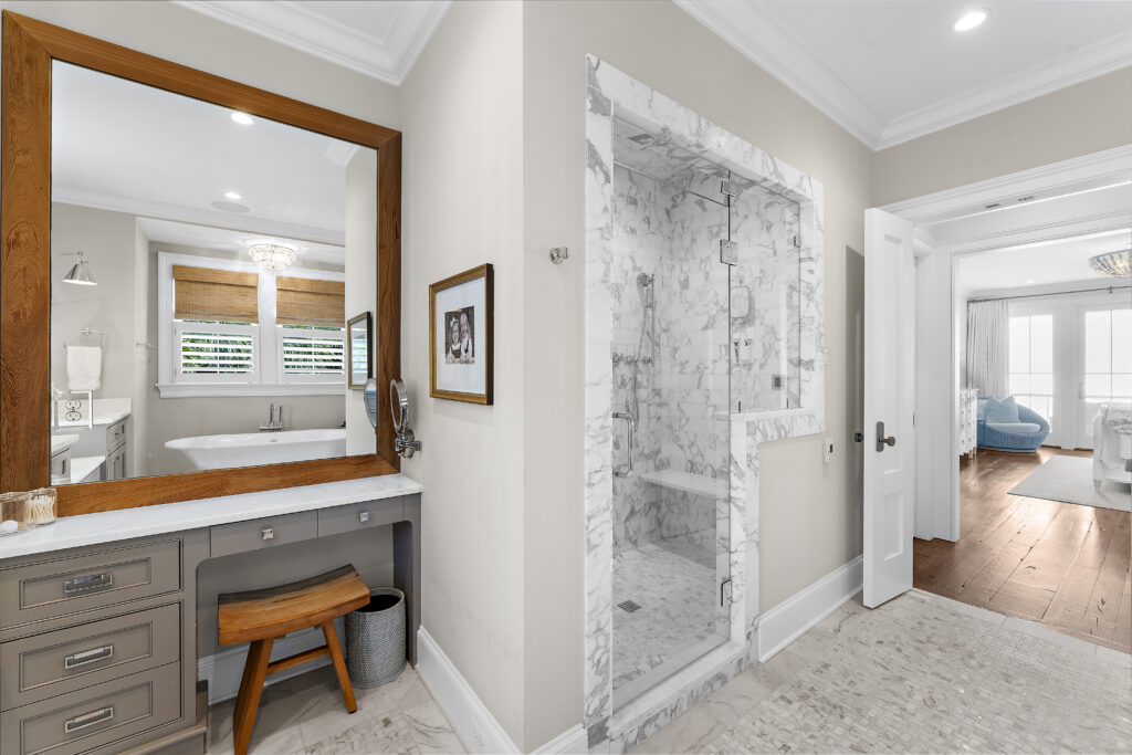 Spacious bathroom with a marble shower reminiscent of April showers, large wood-framed mirror above a vanity with drawers, wooden stool, and a glimpse of a tub by shuttered windows. The adjoining room has hardwood floors and natural light.