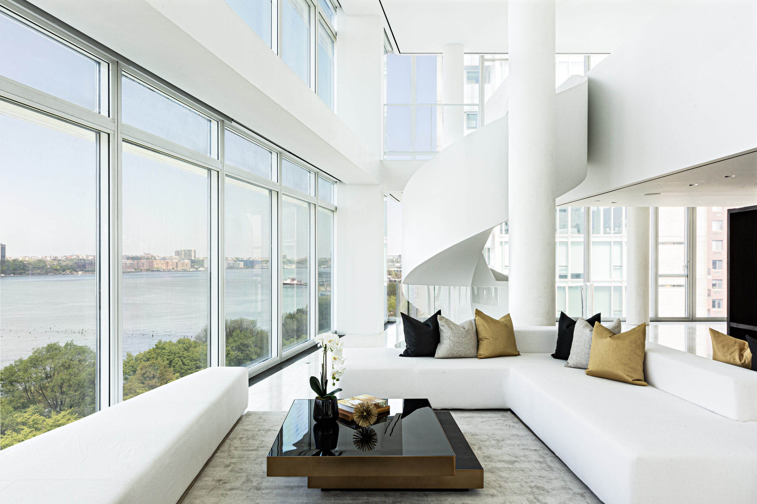 A modern living room with large floor-to-ceiling windows overlooking water, a white curved staircase, stylish sectional sofas, a black coffee table, and minimalistic decor—perfect for an A-list May 2026 gathering.