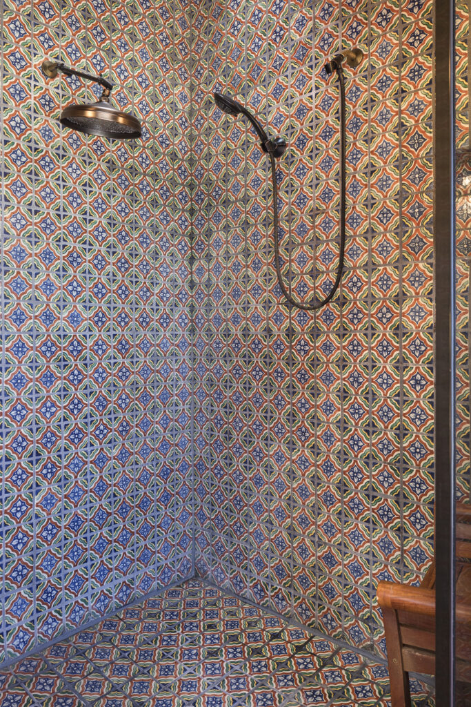 A shower in the primary suite with colorful, intricate geometric patterned tiles on the walls and floor, featuring a large round overhead showerhead and a handheld shower attachment.