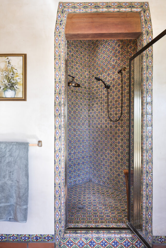 A shower in the primary suite with colorful patterned tiles on the walls, floor, and arched entrance features bronze fixtures. A glass door is partially open; a towel and framed floral art hang on the nearby wall.