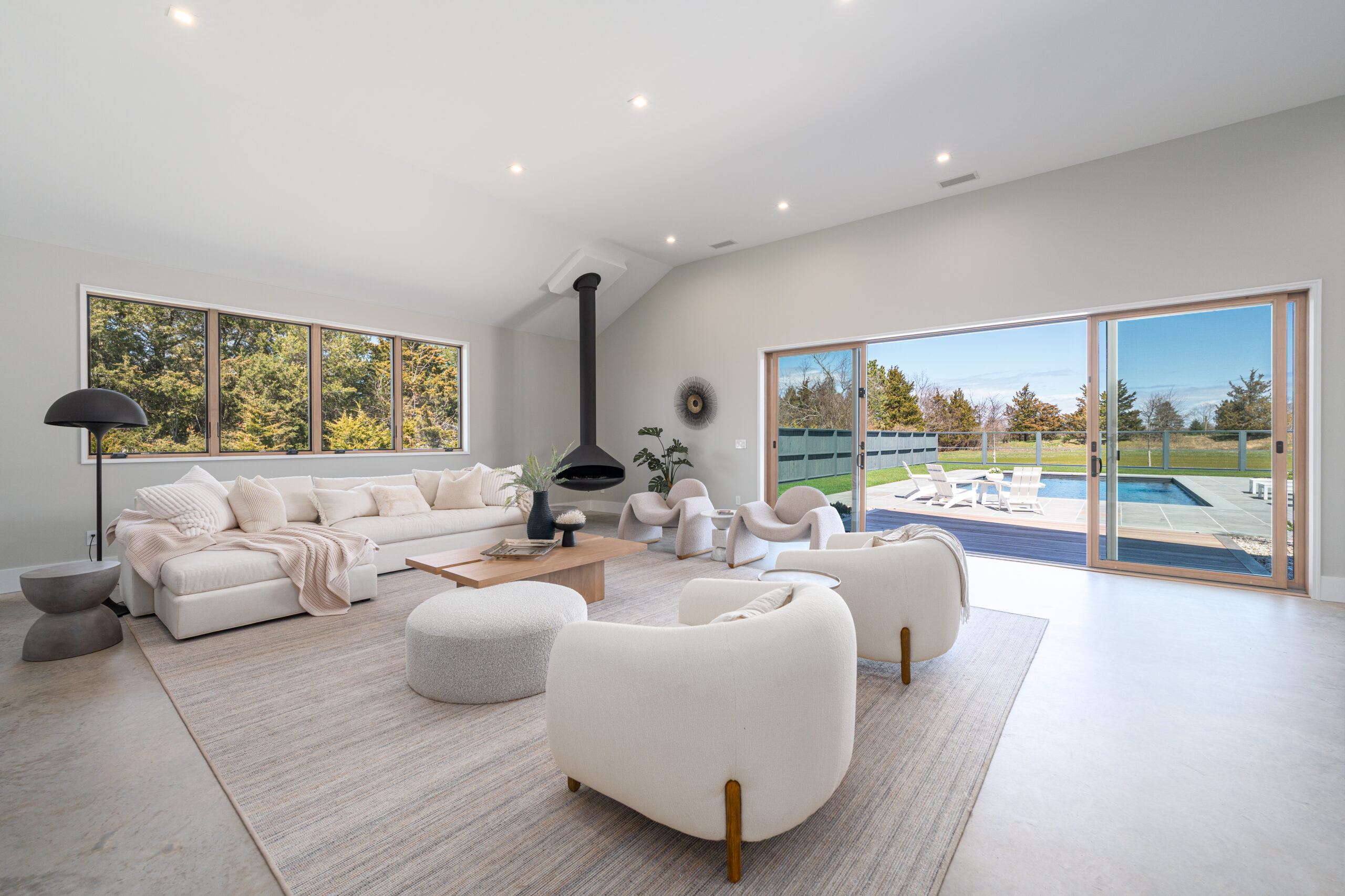 Spacious, modern living room with white furniture, large windows, and sliding glass doors opening to a sunny patio and pool area—an a-list retreat for May 2026. Neutral tones, natural light, and greenery outside create an inviting atmosphere.
