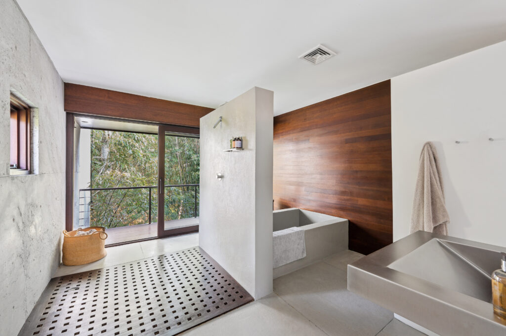 Modern bathroom with minimalist design, featuring a walk-in shower perfect for april showers, concrete tub, large metal sink, wooden accent wall, towel rack, woven basket, and a glass door opening to an outdoor view with greenery.