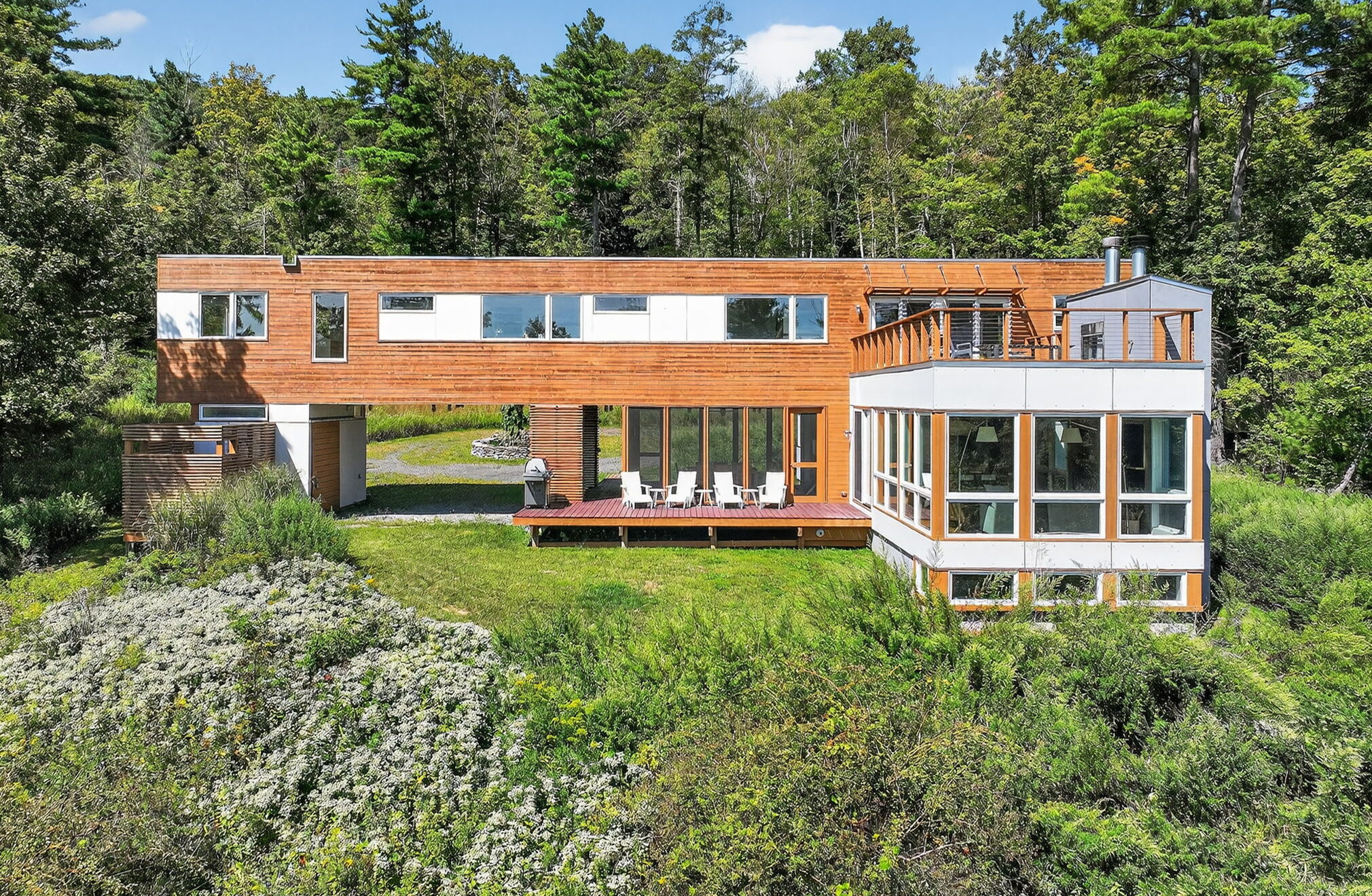 Modern rectangular house with large windows and wooden siding, surrounded by lush greenery and trees. A spacious deck with outdoor seating area overlooks a grassy yard—perfect for an a-list gathering in May 2026.