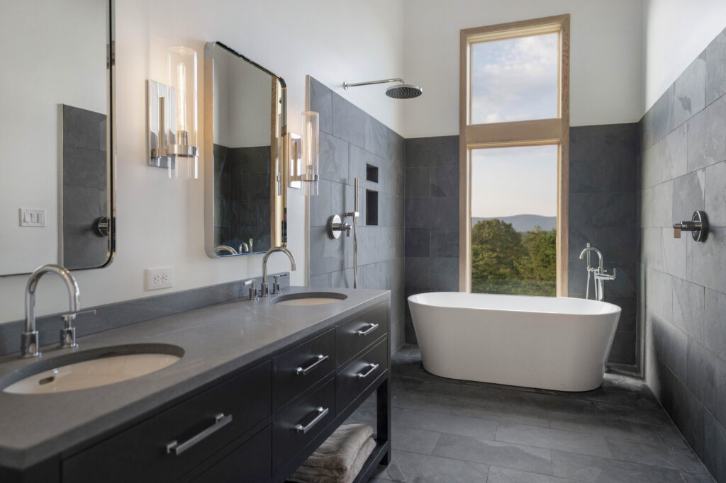 Modern bathroom with double sinks, large mirrors, gray tile walls and floor, a standalone white bathtub beneath a tall window with views of trees and mountains, and a wall-mounted showerhead perfect for relaxing during April showers.