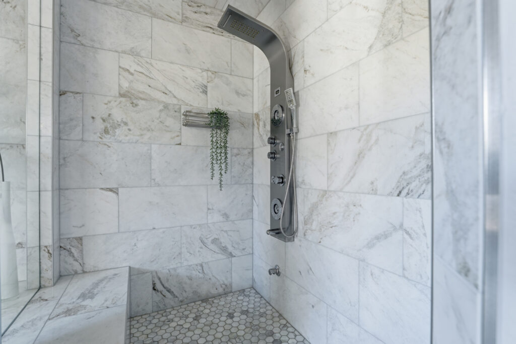 Modern shower with marble tile walls and bench, featuring a sleek silver shower panel with multiple controls and jets. A small shelf holds a trailing green plant, evoking the fresh feel of April showers. The floor is covered with hexagonal tiles.