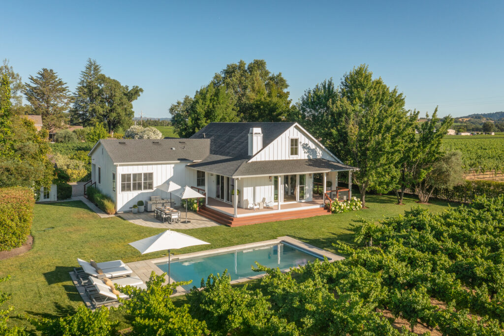A white farmhouse with a large deck overlooks a backyard pool, lounge chairs, and umbrellas, all surrounded by lush green trees, a vineyard, and the scenic views typical of equestrian estates under a clear blue sky.