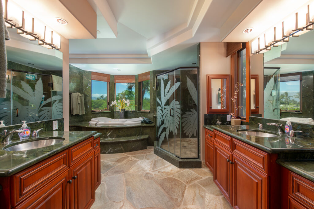 Luxurious bathroom with two wooden vanities, a glass shower perfect for savoring soothing April showers, a tub by large windows, green marble countertops, etched glass leaf panels, stone tile floors, and abundant natural light for a spacious feel.