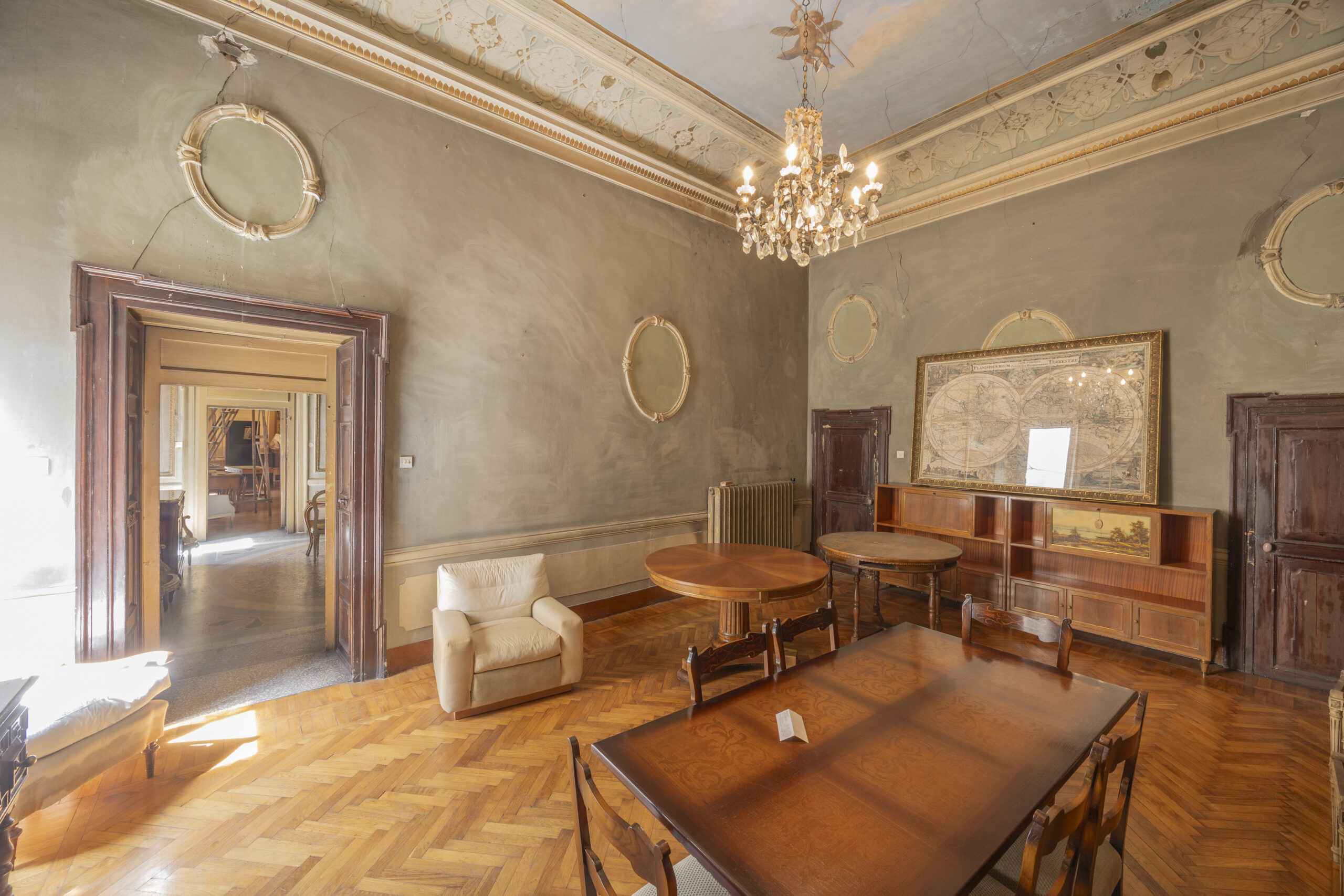 A spacious, elegant room with parquet flooring, ornate ceiling, chandelier, and wooden furniture exudes classic, vintage decor. Framed maps and subtle a-list: May 2026 details add distinction as light streams in from doorways.