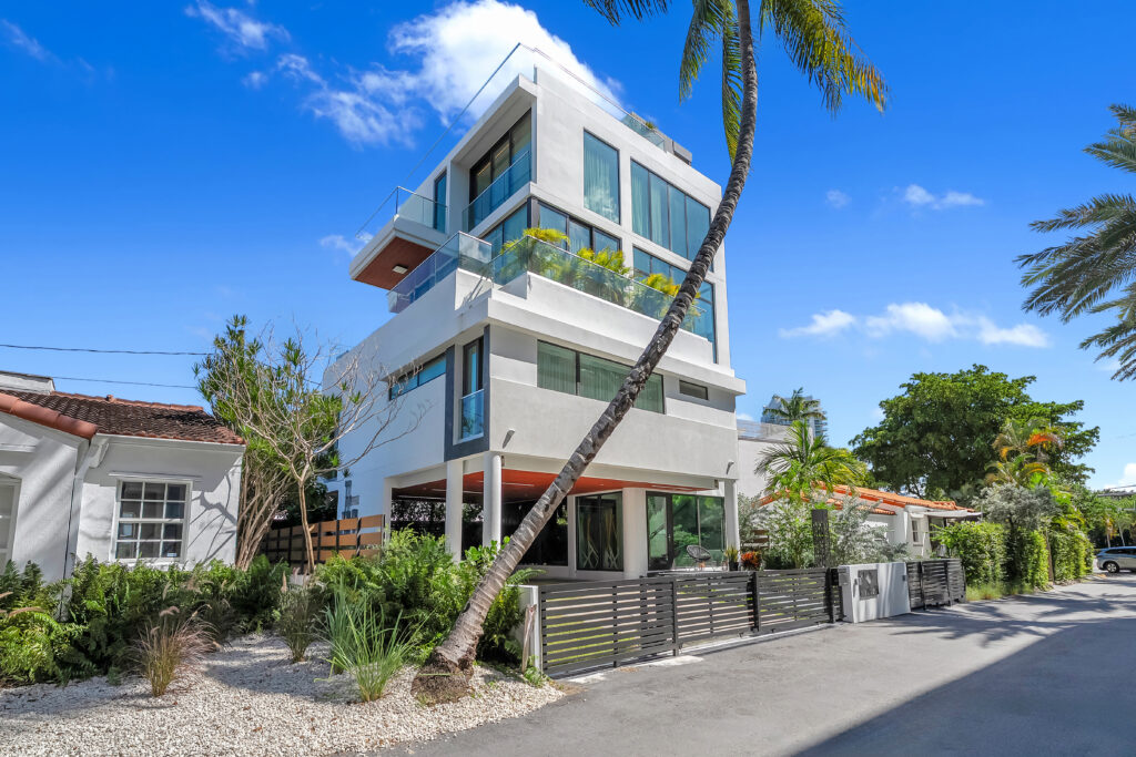 Modern three-story house with large glass windows and balconies, surrounded by tropical plants and palm trees, basking in the calm energy of Taurus season. A sleek metal fence lines the property along a quiet street under a bright blue sky.